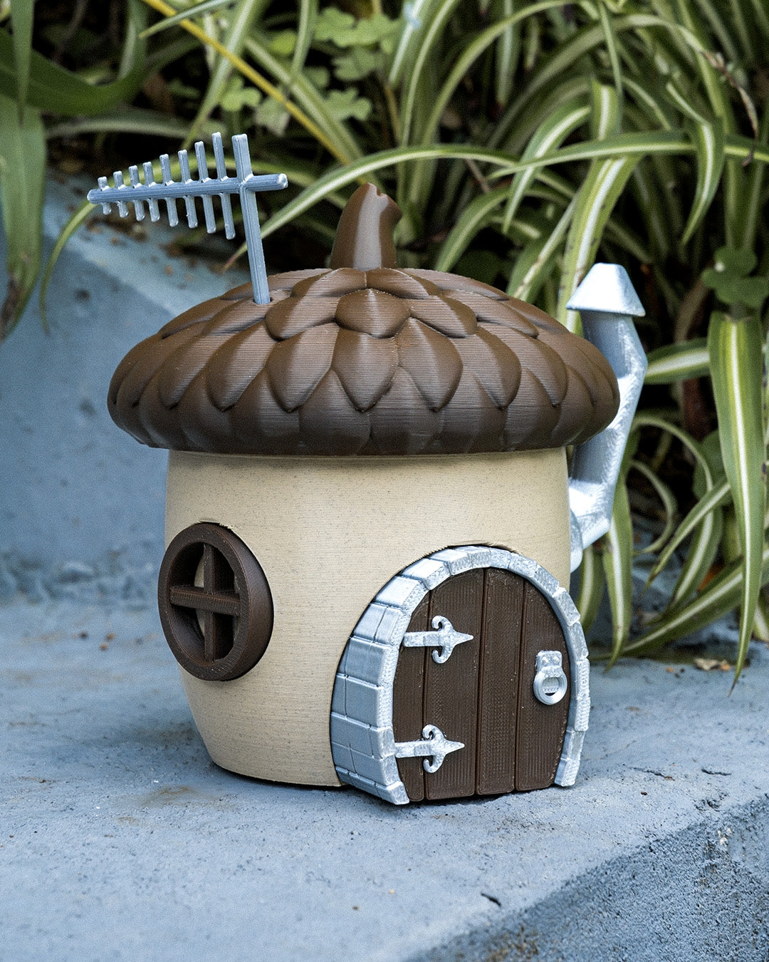 A charming acorn-shaped house decoration featuring a textured brown roof, circular window, wooden door with silver accents, and a chimney. Perfect for gardens, small pet enclosures, or reptile terrariums, set against a natural backdrop of green plants