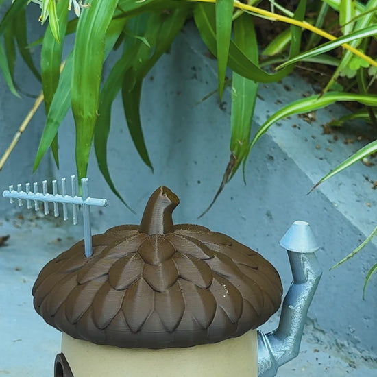 A charming acorn-shaped house decoration featuring a textured brown roof, circular window, wooden door with silver accents, and a chimney. Perfect for gardens, small pet enclosures, or reptile terrariums, set against a natural backdrop of green plants